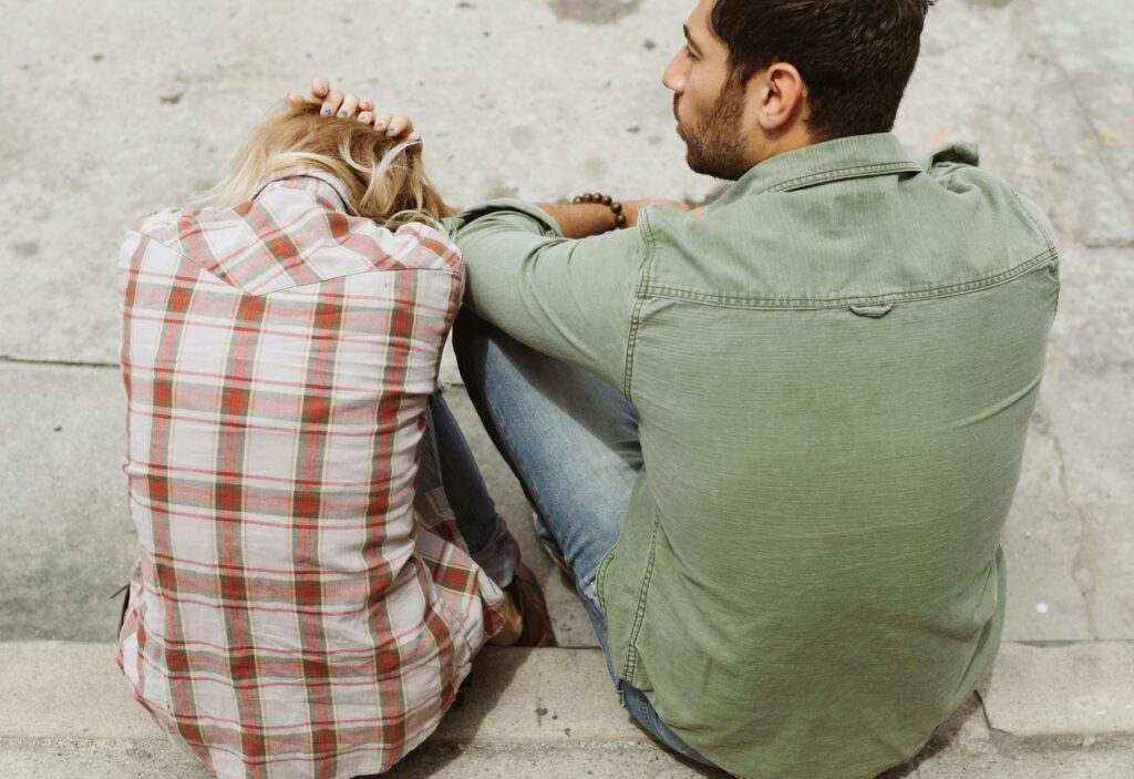 A man and woman sit on steps from behind. The woman, in a plaid shirt, holds her head, appearing upset. The man listens intently, offering support.