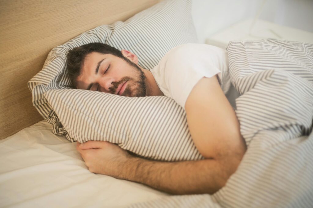A man with a beard peacefully sleeps on a bed, cuddling a striped pillow and covered with a matching blanket. The scene conveys calmness and comfort.