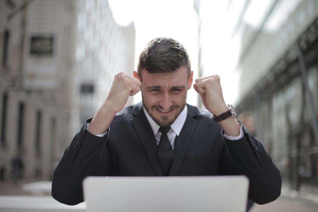 Man in a suit smiling with clenched fists raised in excitement, sitting in front of a laptop, with blurred city buildings in the background.