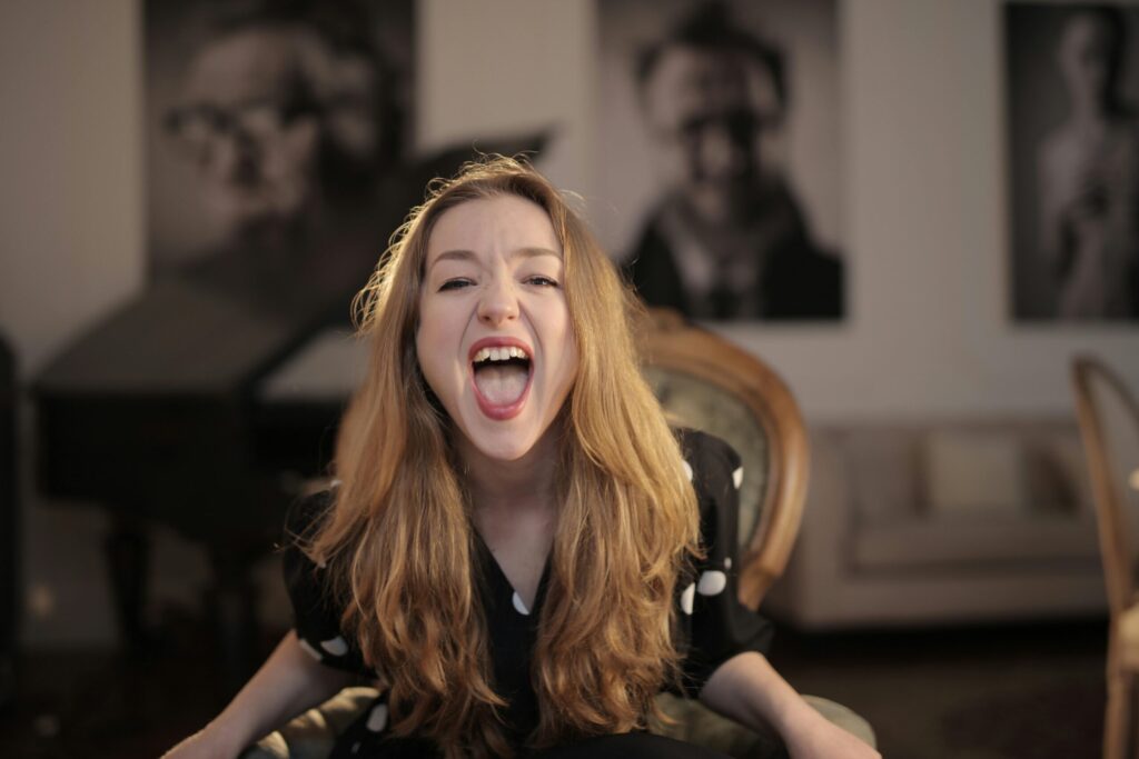 A woman with long blonde hair sits in an armchair, joyfully shouting. The room's soft lighting adds warmth, with blurred portraits decorating the walls.