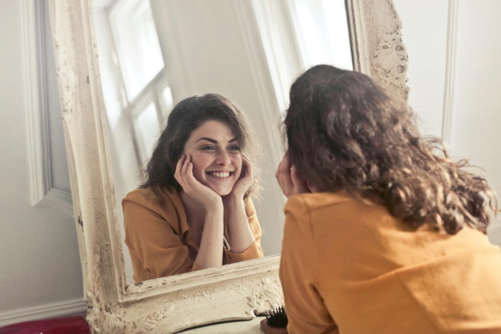 Woman in a mustard shirt smiles at her reflection in an ornate, vintage mirror. Her hands cradle her face, conveying a joyful, relaxed mood.