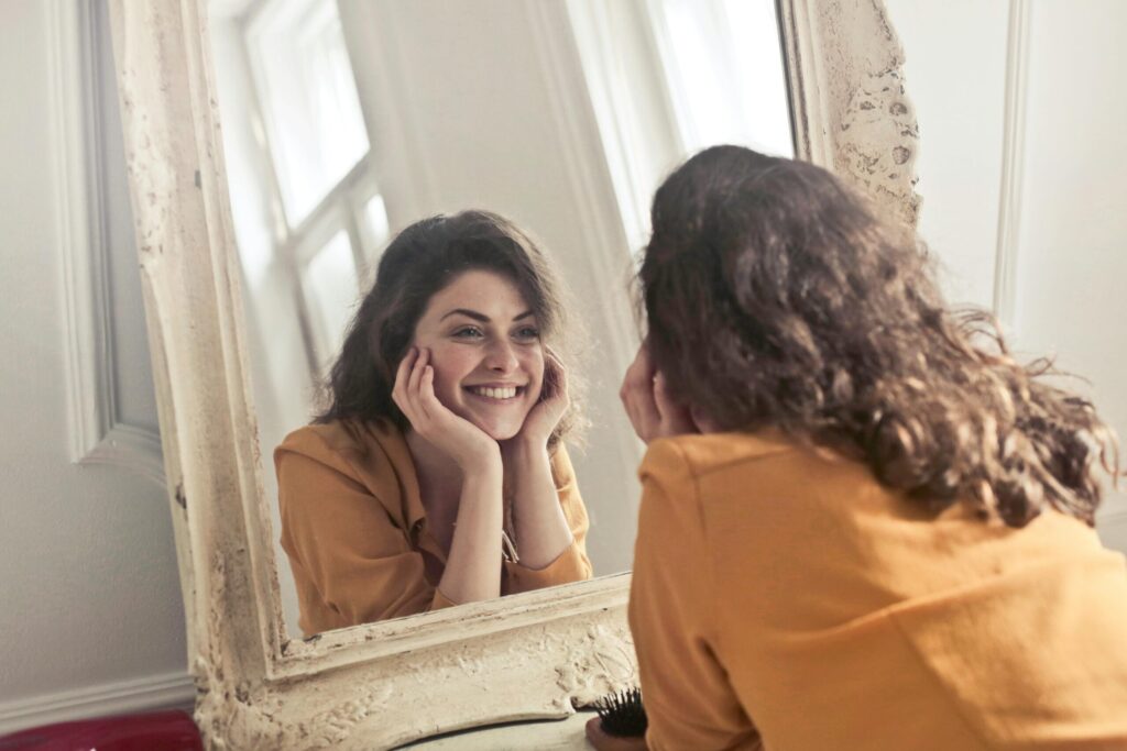 Woman in a mustard shirt smiles at her reflection in an ornate, vintage mirror. Her hands cradle her face, conveying a joyful, relaxed mood.