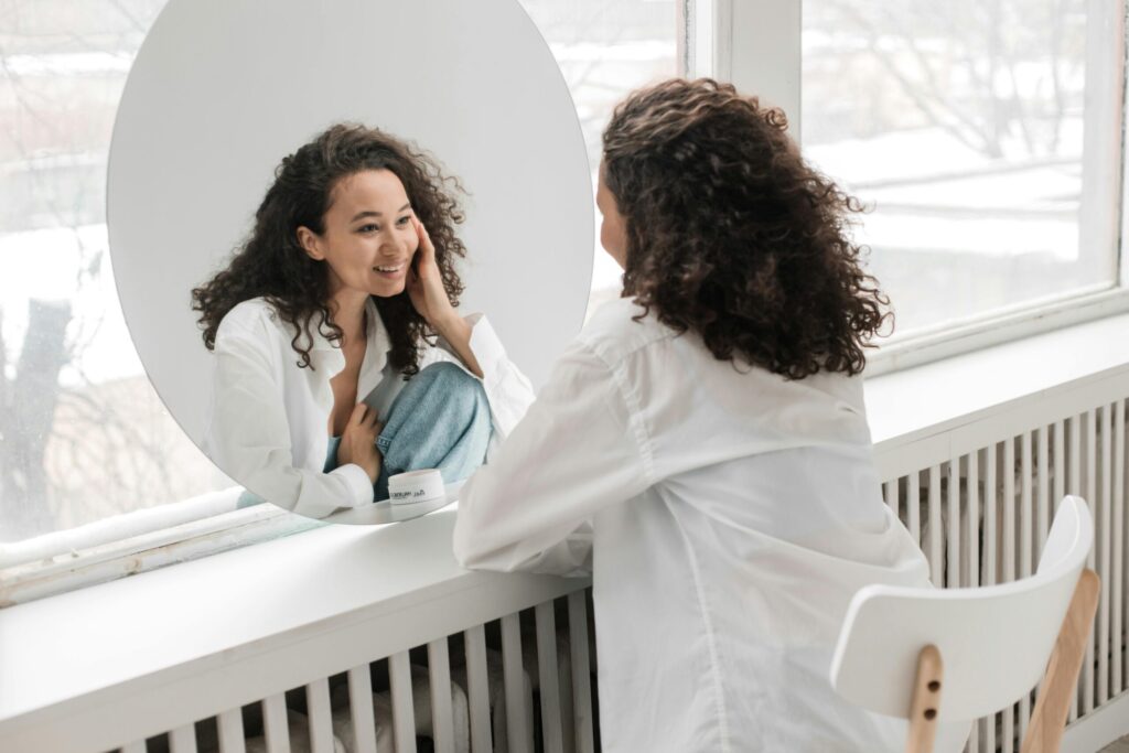 Woman with curly hair, wearing a white shirt, smiles at her reflection in a round mirror. She sits on a white chair in a bright room, conveying a joyful mood.