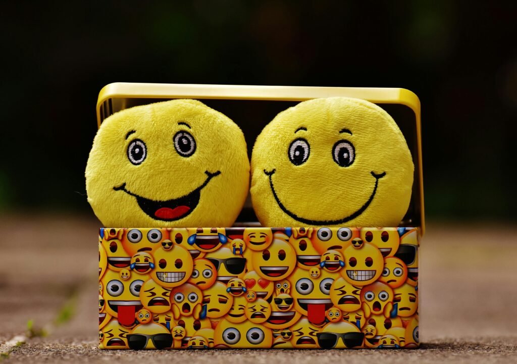 Two plush smiley faces sit in a colorful box adorned with various emoji designs. The setting is playful, conveying happiness and fun.