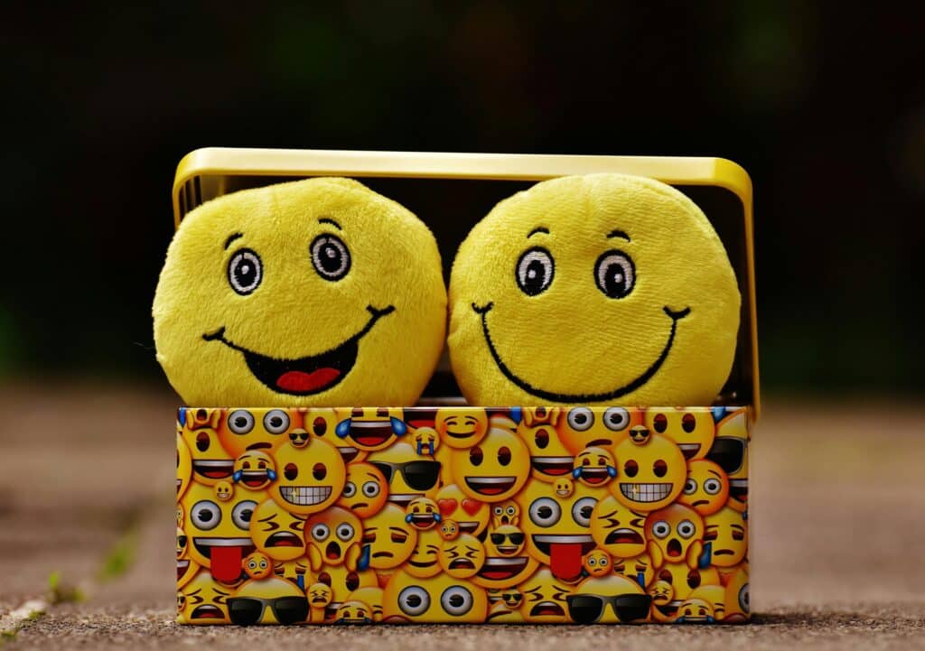 Two plush smiley faces sit in a colorful box adorned with various emoji designs. The setting is playful, conveying happiness and fun.