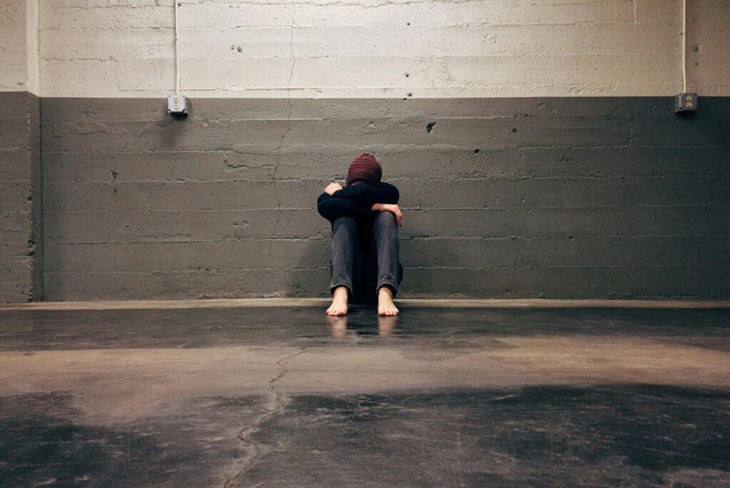 Person sitting barefoot on a concrete floor against a gray, somber wall, hugging knees with head down. The scene conveys loneliness and contemplation.