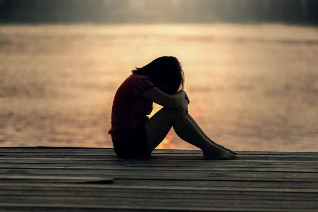 A silhouette of a seated person on a wooden dock at sunset, hugging their knees. The warm, golden glow of the water sets a contemplative mood.