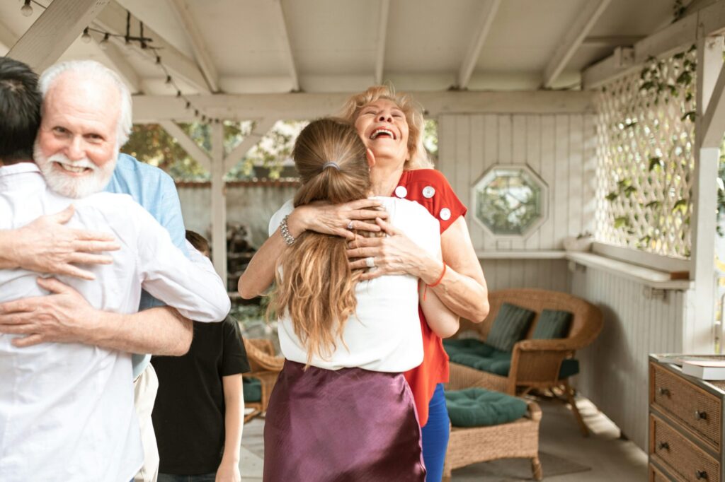 Elderly woman joyfully hugs a younger person in a wicker-furnished patio. A smiling older man hugs another person nearby, creating a warm, cheerful atmosphere.