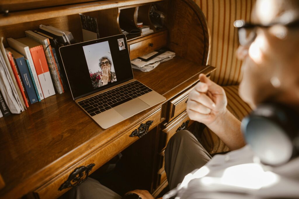 A man wearing headphones is at a wooden desk, video chatting on a laptop with an elderly woman who appears happy, blowing a kiss. Books are visible.