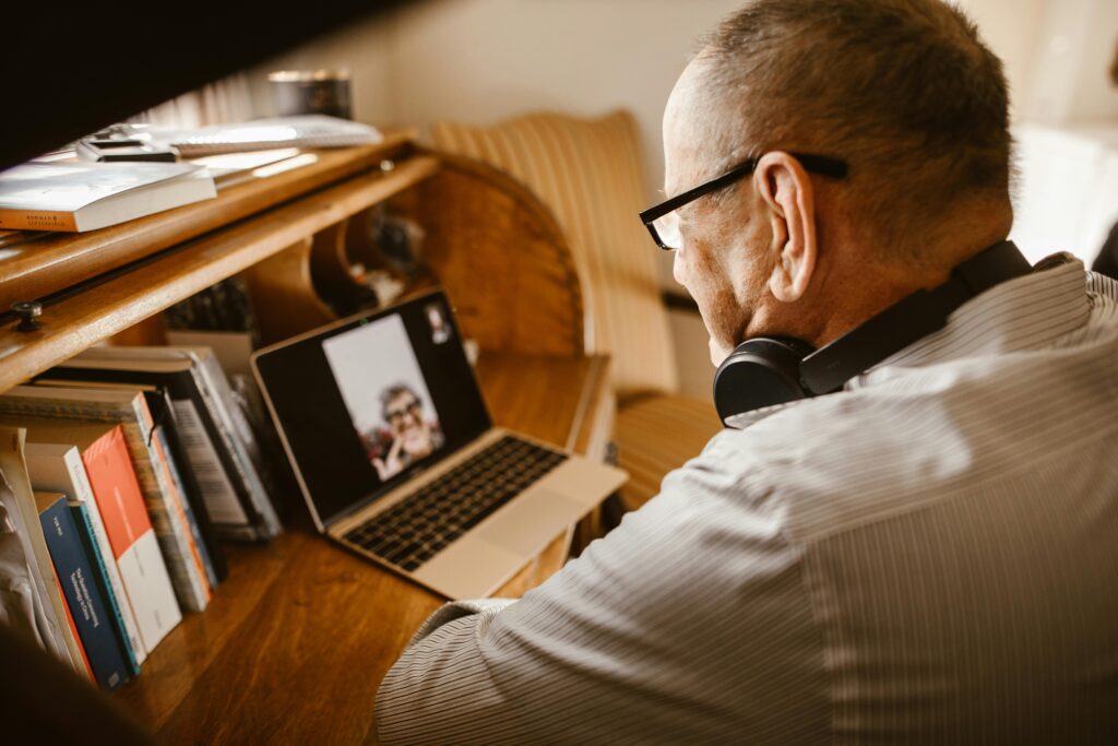 A man wearing headphones and glasses is video chatting on a laptop in a home office. Books and a notepad are on a wooden desk, creating a cozy atmosphere.