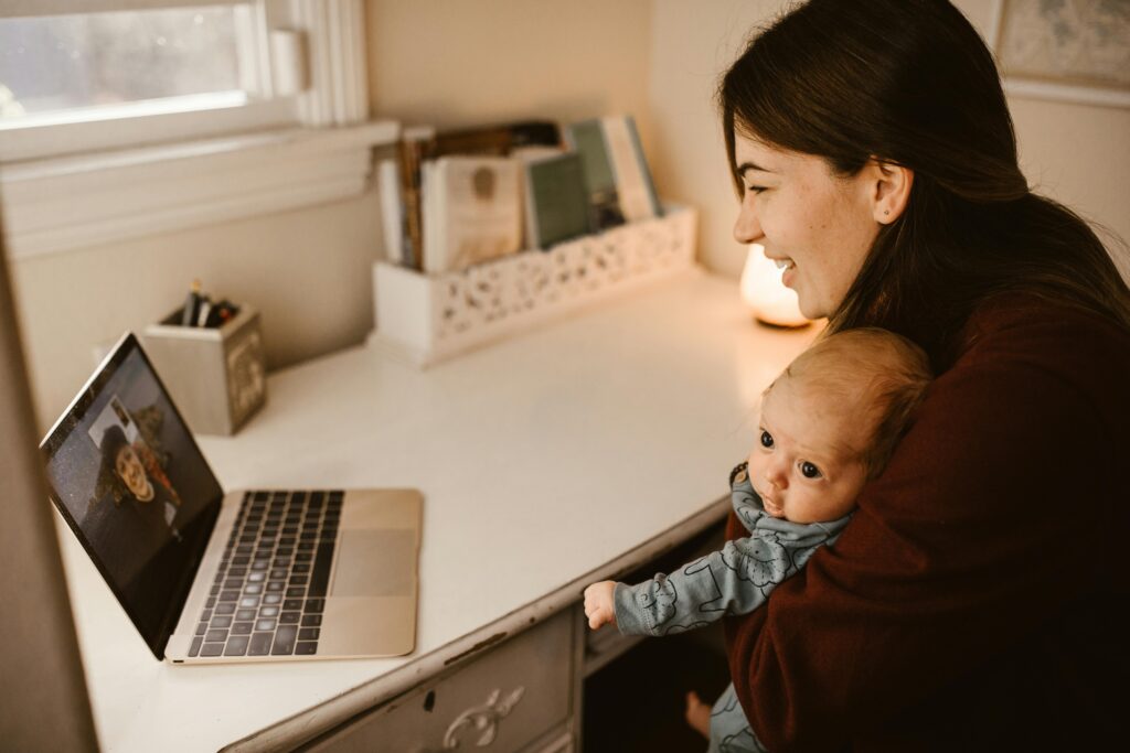 A woman smiles while holding a baby and video chatting on a laptop. The cozy room and warm lighting create a joyful, intimate atmosphere.