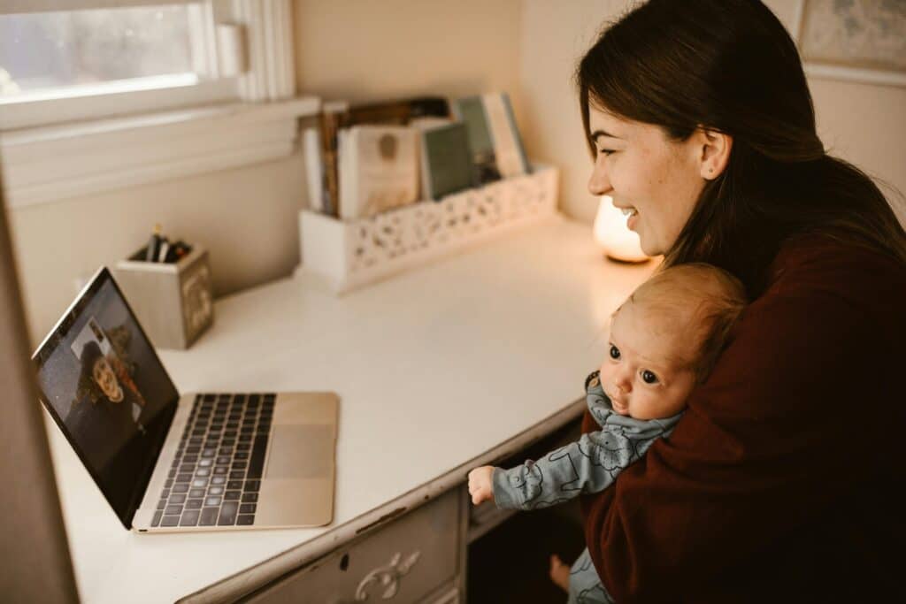A woman smiles while holding a baby and video chatting on a laptop. The cozy room and warm lighting create a joyful, intimate atmosphere.