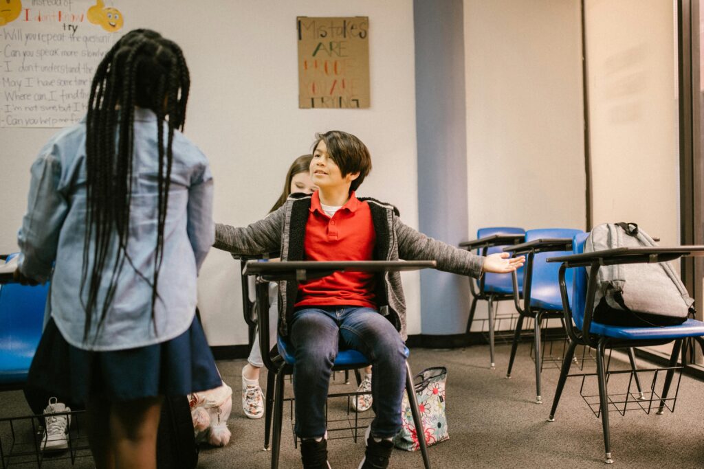 A child in a red shirt sits at a school desk, arms spread wide, facing another child standing. Blue chairs and motivational posters are visible, conveying a learning environment.