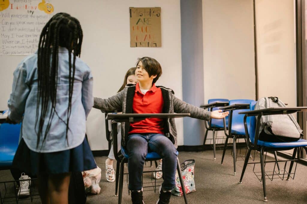 A child in a red shirt sits at a school desk, arms spread wide, facing another child standing. Blue chairs and motivational posters are visible, conveying a learning environment.