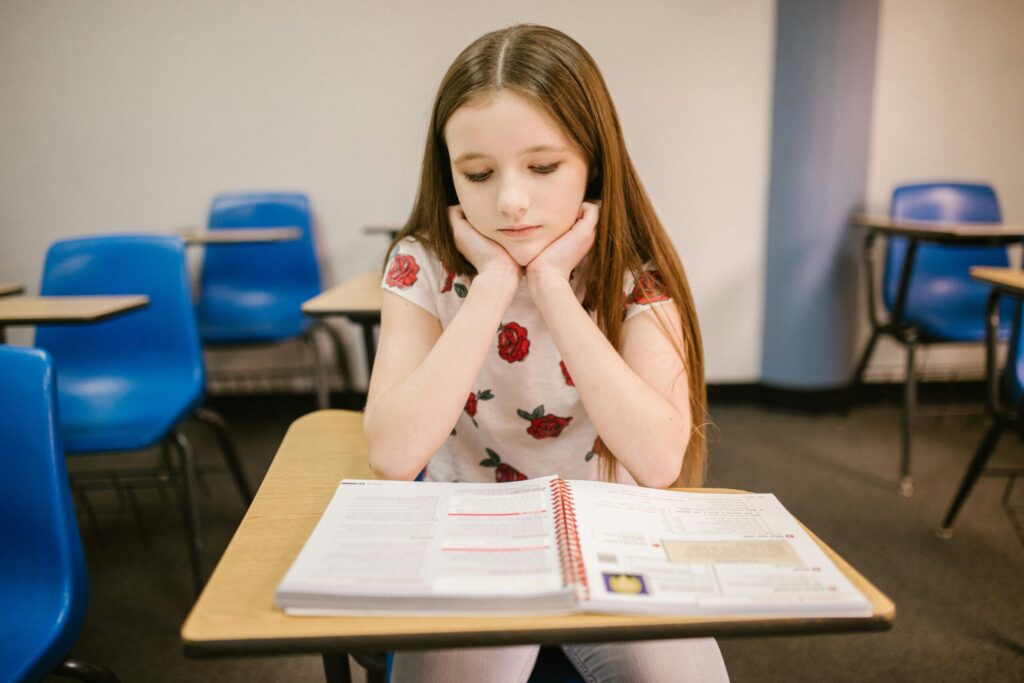 A young girl sits at a school desk, reading a textbook with a thoughtful expression. She wears a floral-patterned shirt. Blue chairs are visible in the background.