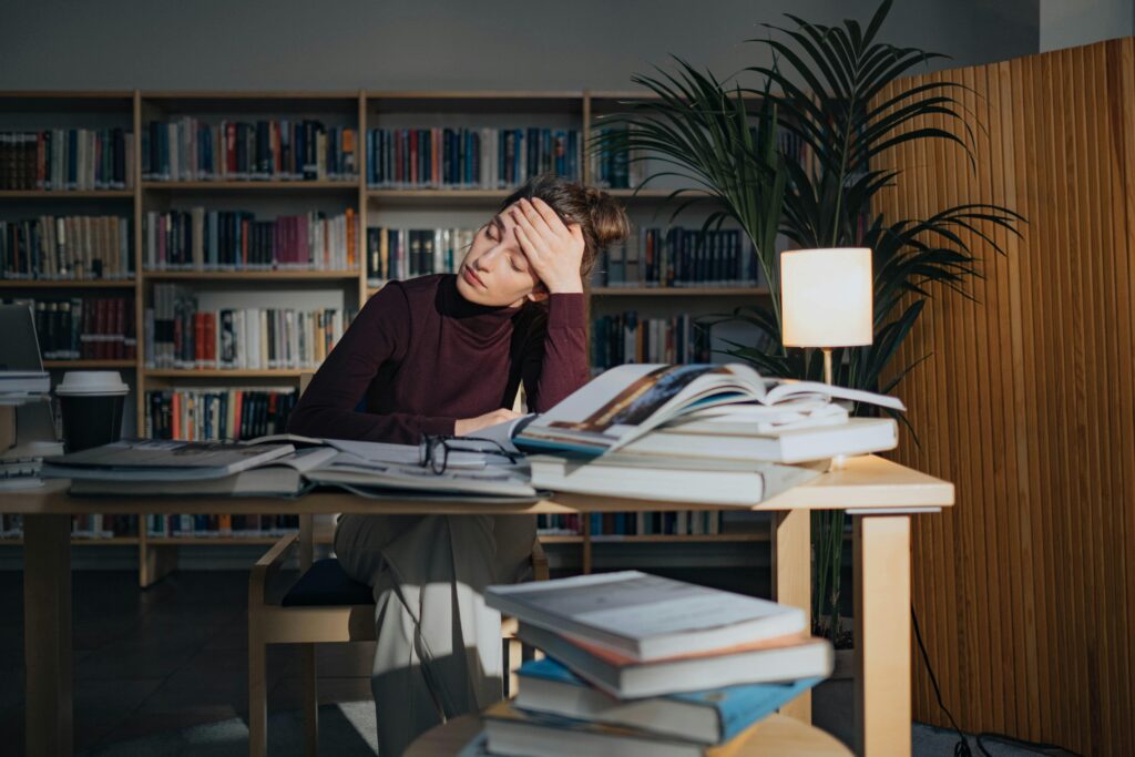 A tired woman sits at a library table, head resting on hand, surrounded by open books and papers. Soft lamp light and bookshelves create a studious ambiance.