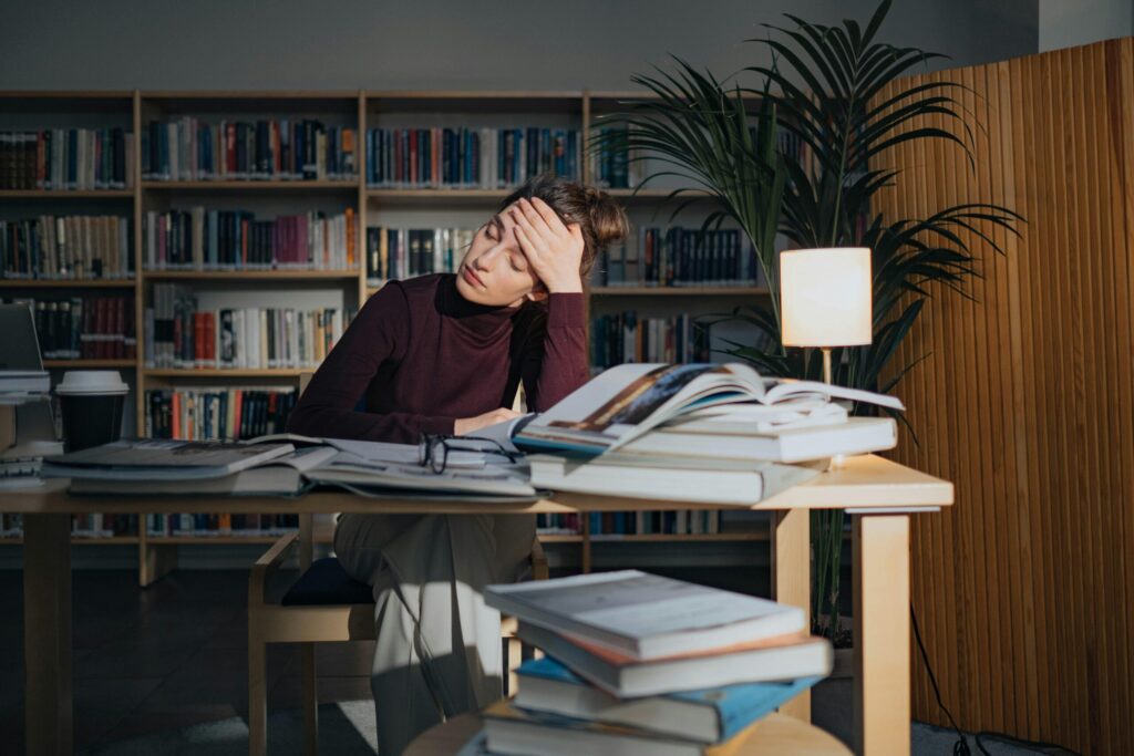 A tired woman sits at a library table, head resting on hand, surrounded by open books and papers. Soft lamp light and bookshelves create a studious ambiance.