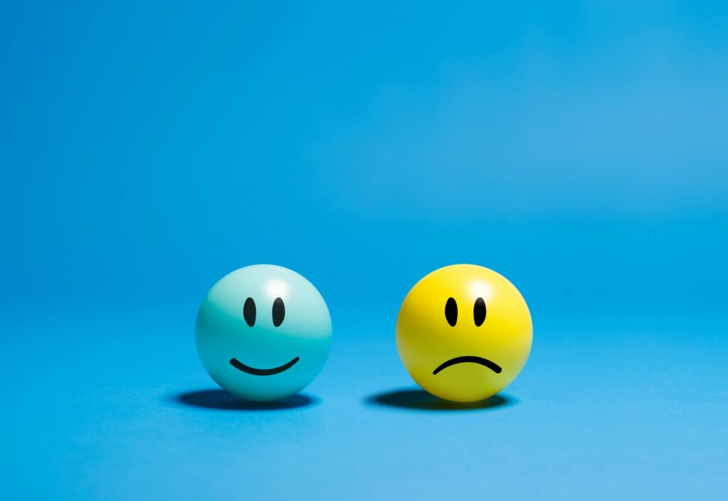 Two balls on a blue background; a light blue ball with a happy face and a yellow ball with a sad face, symbolizing contrasting emotions.
