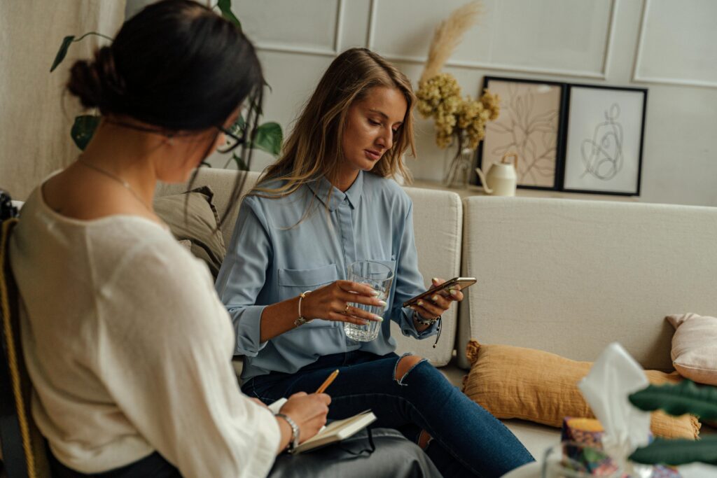 Two women in casual clothing sit on a sofa, engaged in conversation. One checks her phone and holds a glass of water. The space feels cozy and relaxed.