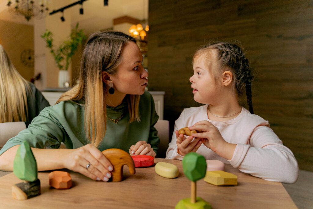 A woman and a young girl with Down syndrome engage in play with colorful wooden toys at a table. The scene is warm, conveying care and connection.