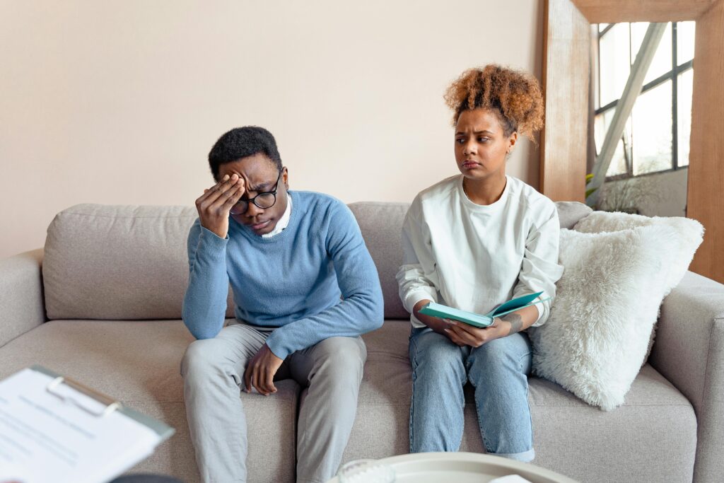 A worried couple sits on a beige sofa during a therapy session. The man looks distressed with a hand on his forehead; the woman holds a notebook.