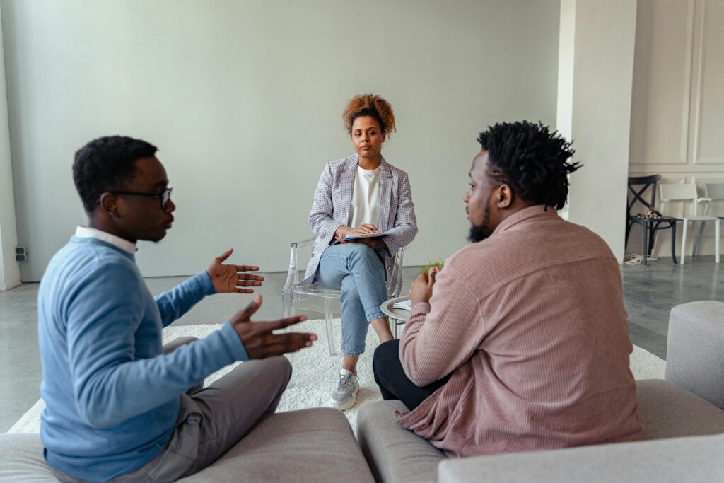 Three people engage in a discussion in a minimal room. Two men sit on a couch gesturing, while a woman listens attentively across from them.