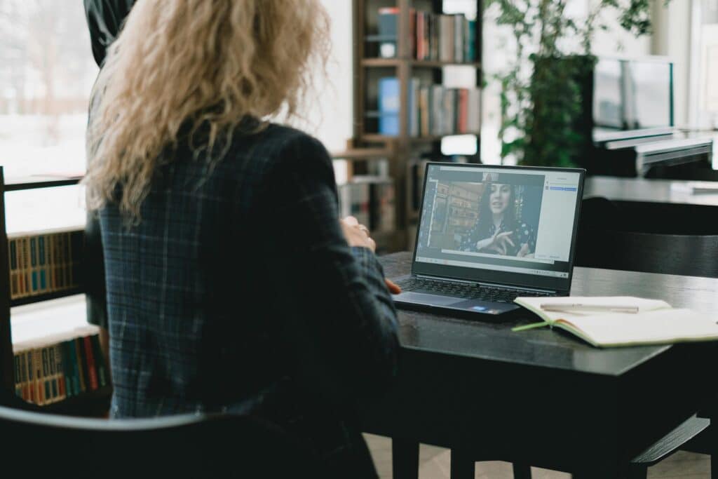 A woman with curly hair uses a laptop in a library setting. The screen shows a video call. An open notebook and pen are on the table.
