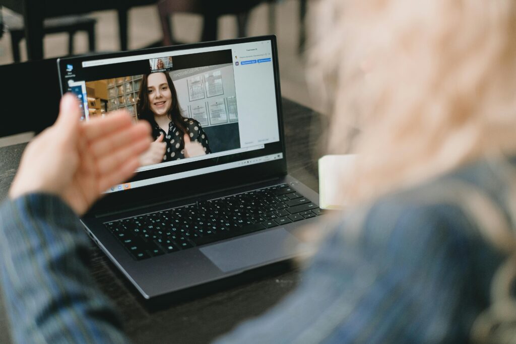 A person in a striped shirt is on a video call, watching someone on a laptop screen giving a thumbs-up. The mood is positive and engaging.