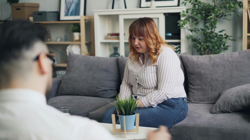 A woman with red hair sits pensively on a gray couch, facing a man with glasses in an office setting. A plant on the table creates a calming mood.