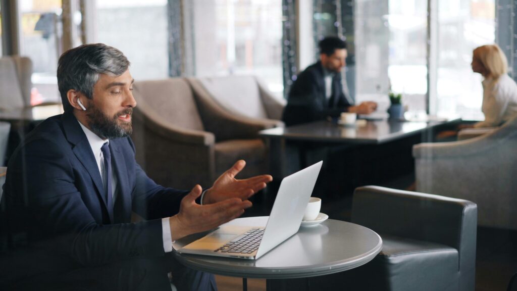 A man in a suit uses a laptop, gesturing during a video call in a café. Two blurred figures in the background create a busy and professional atmosphere.