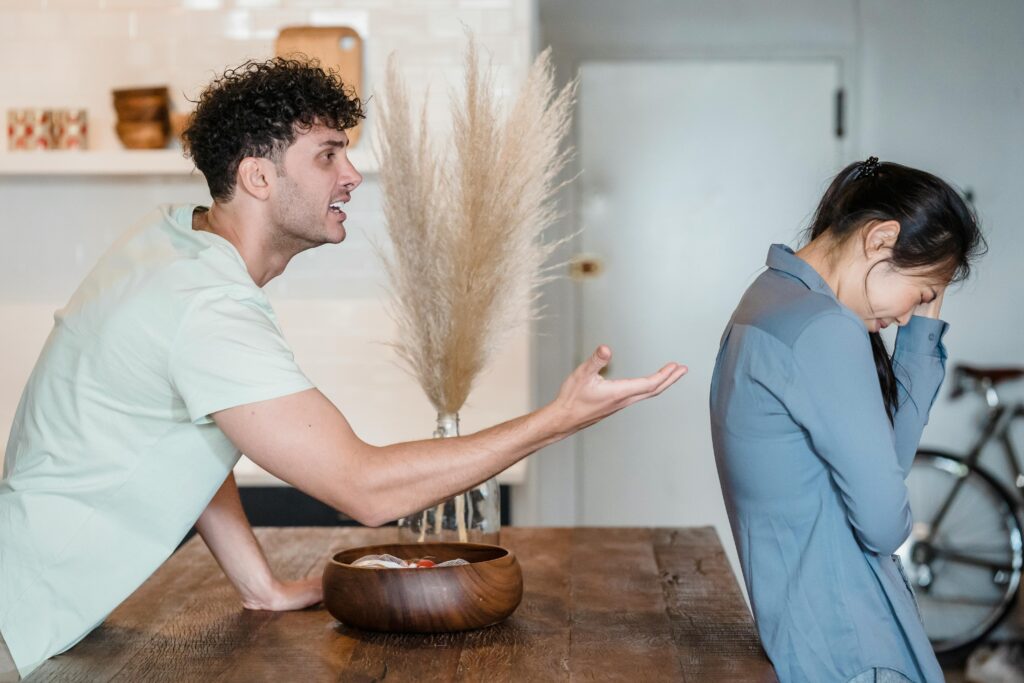 Man with curly hair appears upset, gesturing while speaking to a woman in blue who looks distressed, covering her face. Tense indoor setting.
