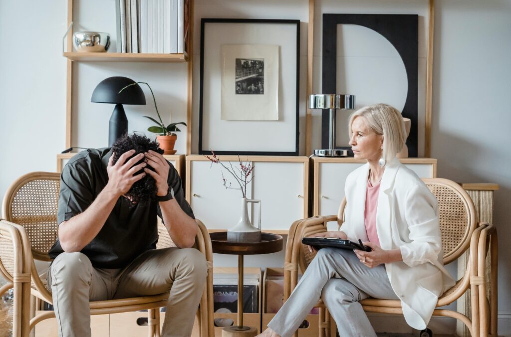 A woman in a white blazer sits attentively with a tablet, facing a distressed man holding his head. They are in a cozy office space with art and plants.