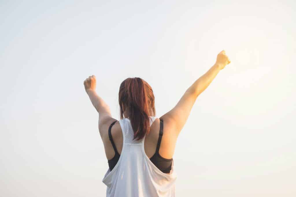 A woman with her back to the camera raises her arms triumphantly towards a bright sky, conveying a sense of achievement and freedom.