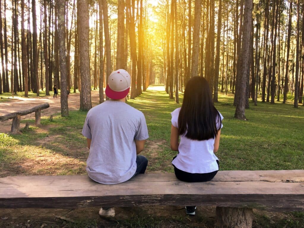 A man and a woman sit on a wooden bench in a sunlit forest, back facing the camera. Tall trees surround them, evoking a peaceful, serene atmosphere.