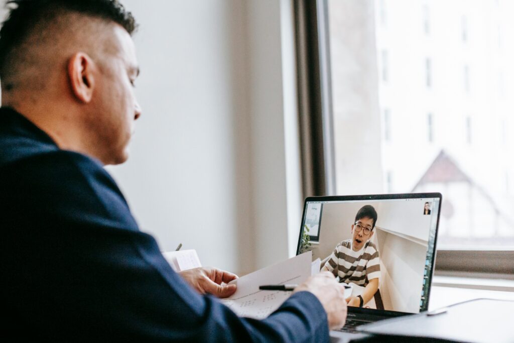 A man in a navy blazer sits at a table, engaged in a video call on a laptop with a young person in striped clothes. The setting feels focused and attentive.