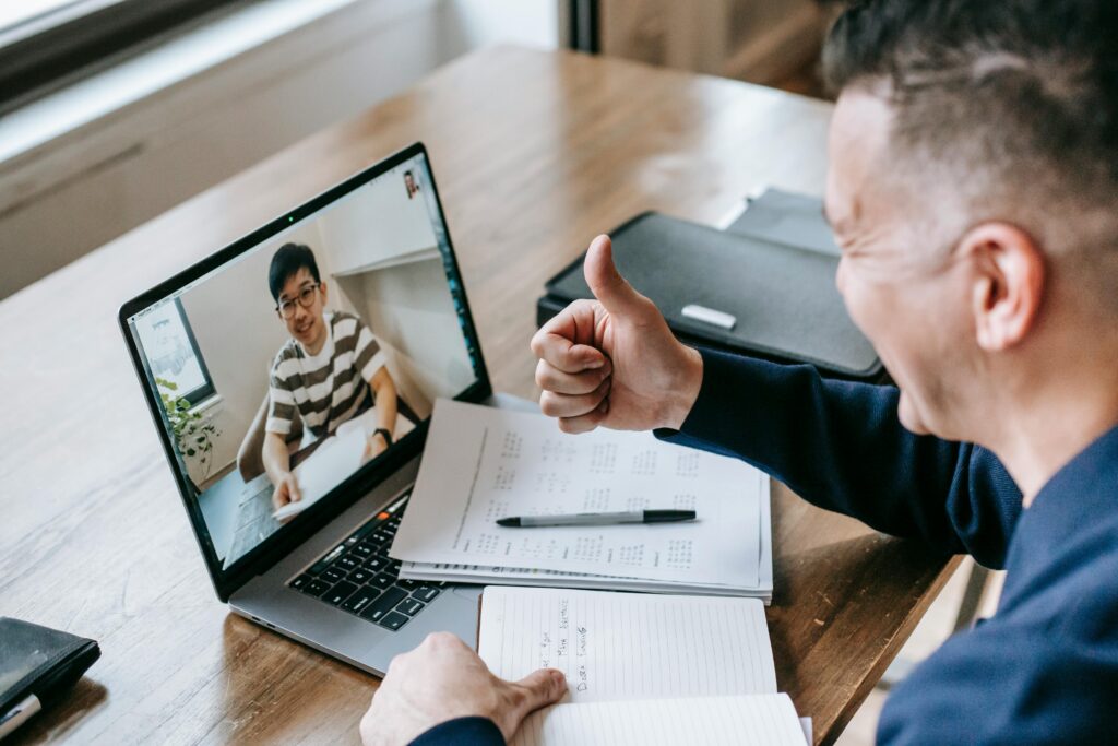 Man giving thumbs-up during a video call on a laptop, smiling warmly. Desk holds papers, pen, and notebook, creating a productive and positive atmosphere.