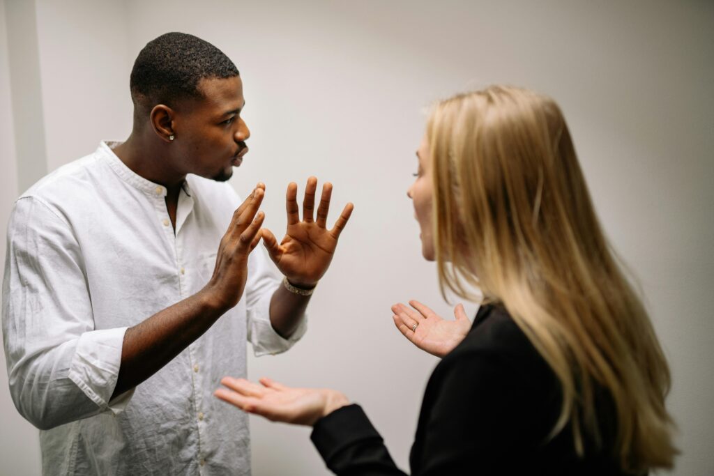 Two people in heated discussion. A man in a white shirt gestures with both hands, while a woman in a black outfit responds, expressing disagreement.