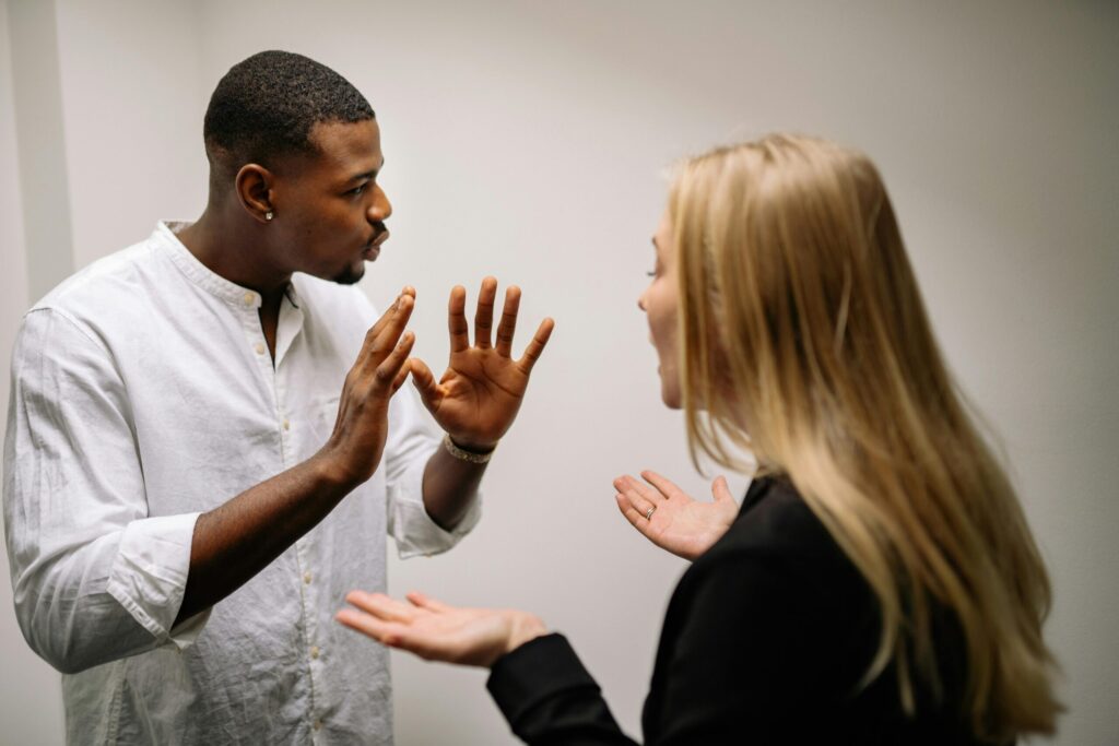 Two people in heated discussion. A man in a white shirt gestures with both hands, while a woman in a black outfit responds, expressing disagreement.