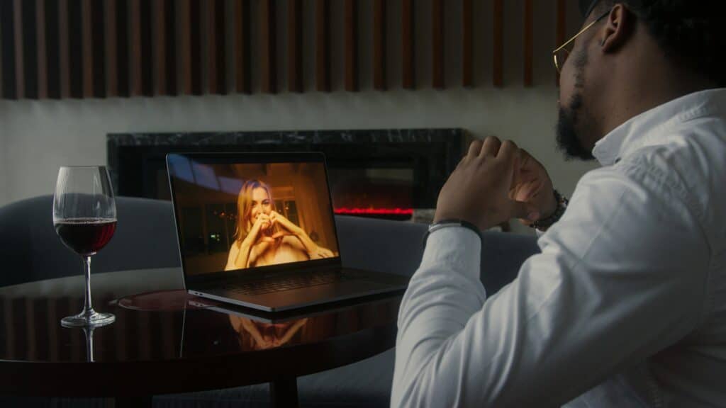 A person in a white shirt sits at a table, video chatting on a laptop with a woman making a heart gesture. A glass of red wine is beside the laptop.