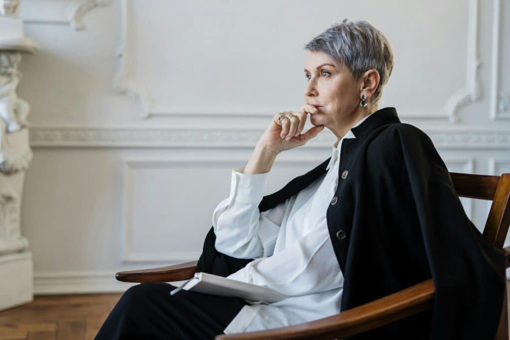 A thoughtful individual in a stylish outfit sits in a wooden chair, holding a notebook in an elegantly decorated room.