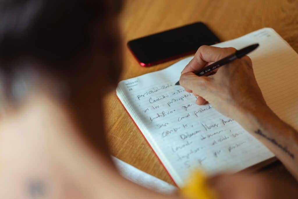 A person writes in a notebook with a black pen, notes visible on the page, while a smartphone rests nearby on a wooden surface.