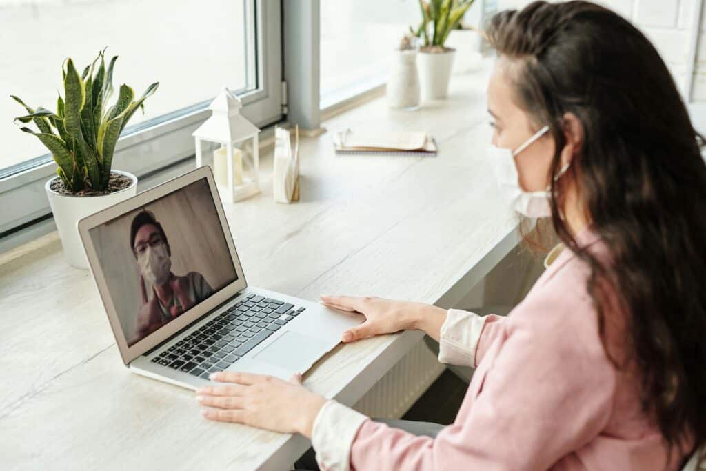 A person in a light pink shirt sits at a desk, engaging in a video call on a laptop, surrounded by indoor plants.