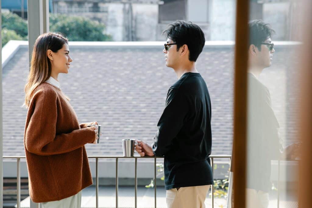 A woman in a cozy brown sweater and a man in a black shirt stand on a balcony, engaged in conversation with cups in hand.