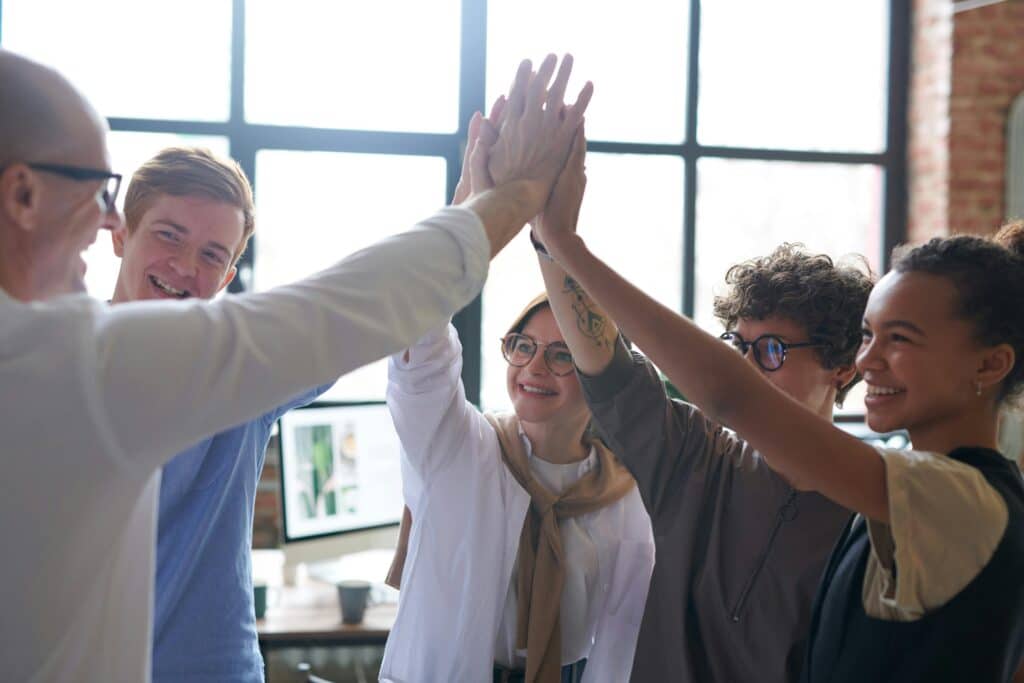 A group of diverse individuals celebrate together by raising their hands in unity, with a bright workspace in the background.