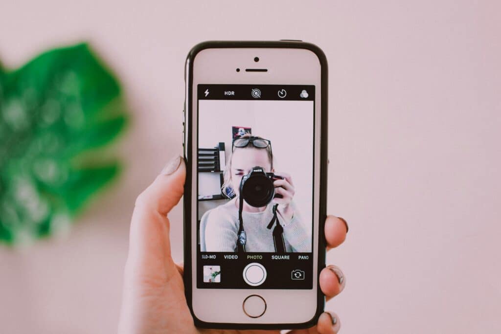 A hand holds a smartphone displaying a selfie, with a blurred face, against a soft pink wall and a green plant in the background.