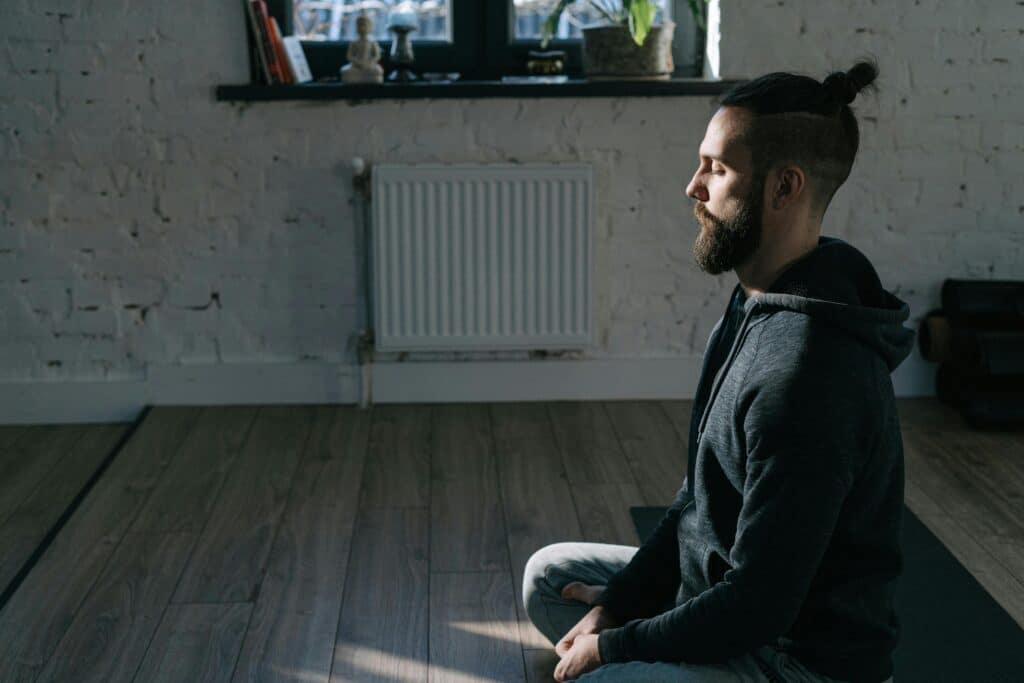 A person in a dark hoodie sits cross-legged on a yoga mat in a serene, softly lit room with a textured white wall.