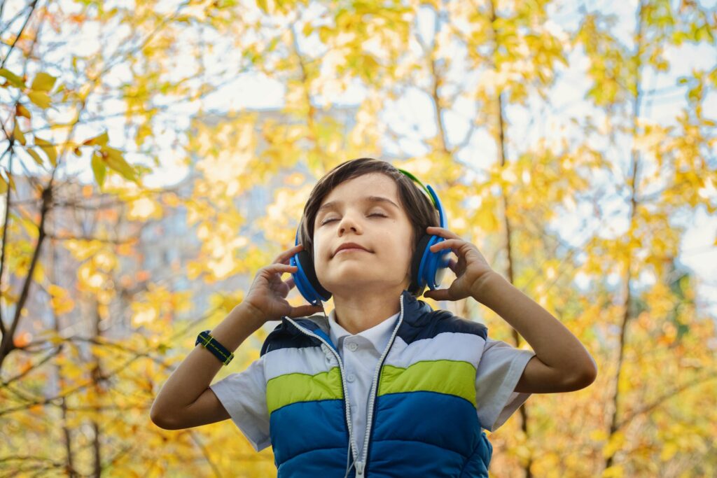 A child wearing blue headphones stands outdoors amidst trees with vibrant autumn leaves, enjoying music in a sunny environment.