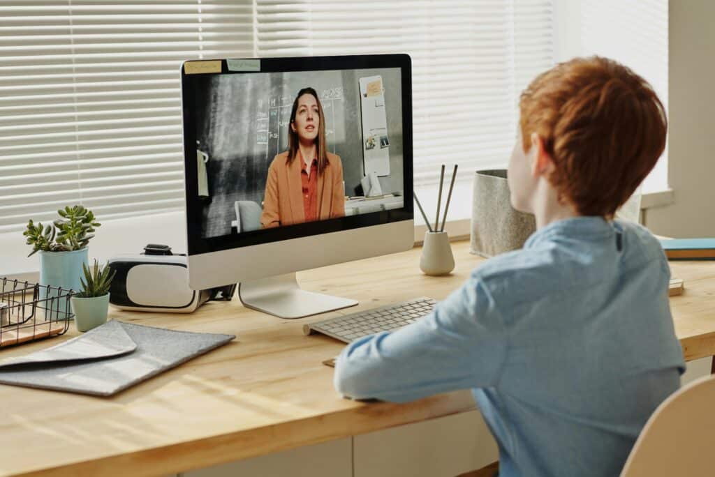 A child observes a virtual class on a desktop computer, with a potted plant and stationery on a wooden desk in a well-lit room.