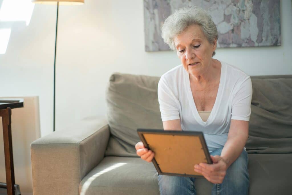 An elderly woman sitting on a couch, holding a picture frame, surrounded by a cozy living room setting with soft lighting.
