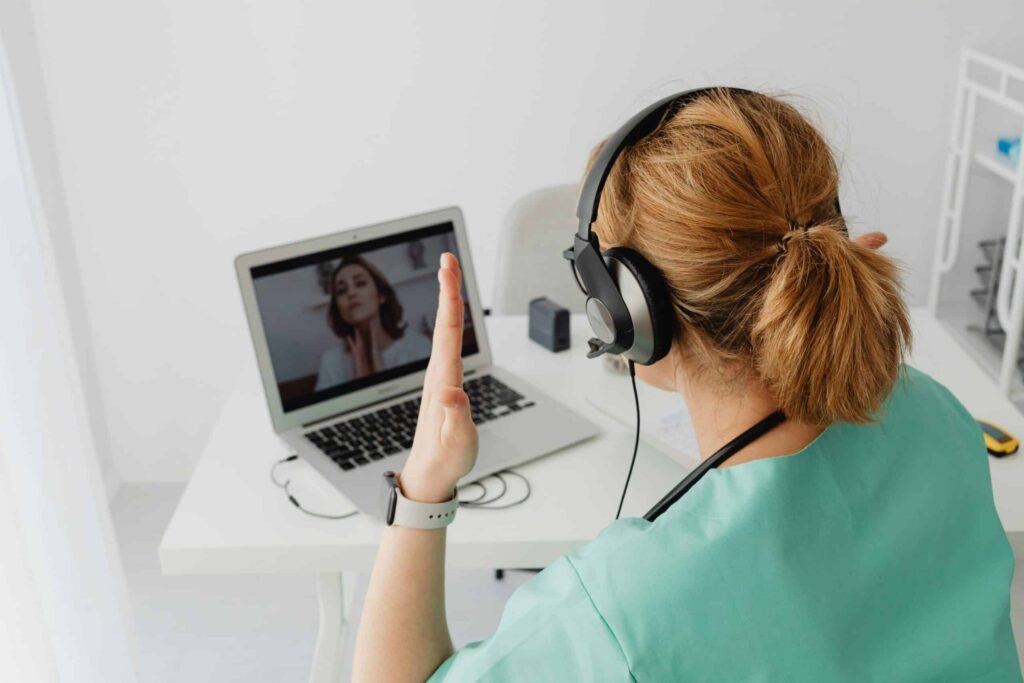 A person in a teal shirt wearing headphones engages in a video call on a laptop, raising a hand to greet the other participant.